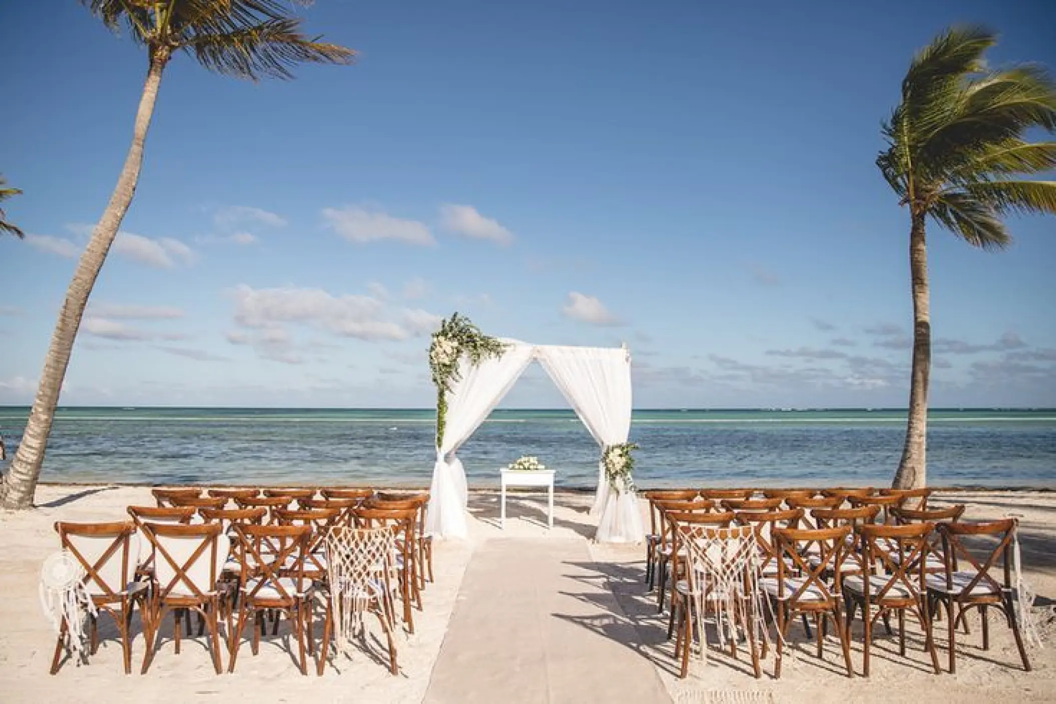 Beachfront wedding ceremony with white draped arch and wooden chairs at Secrets Cap Cana, Punta Cana
