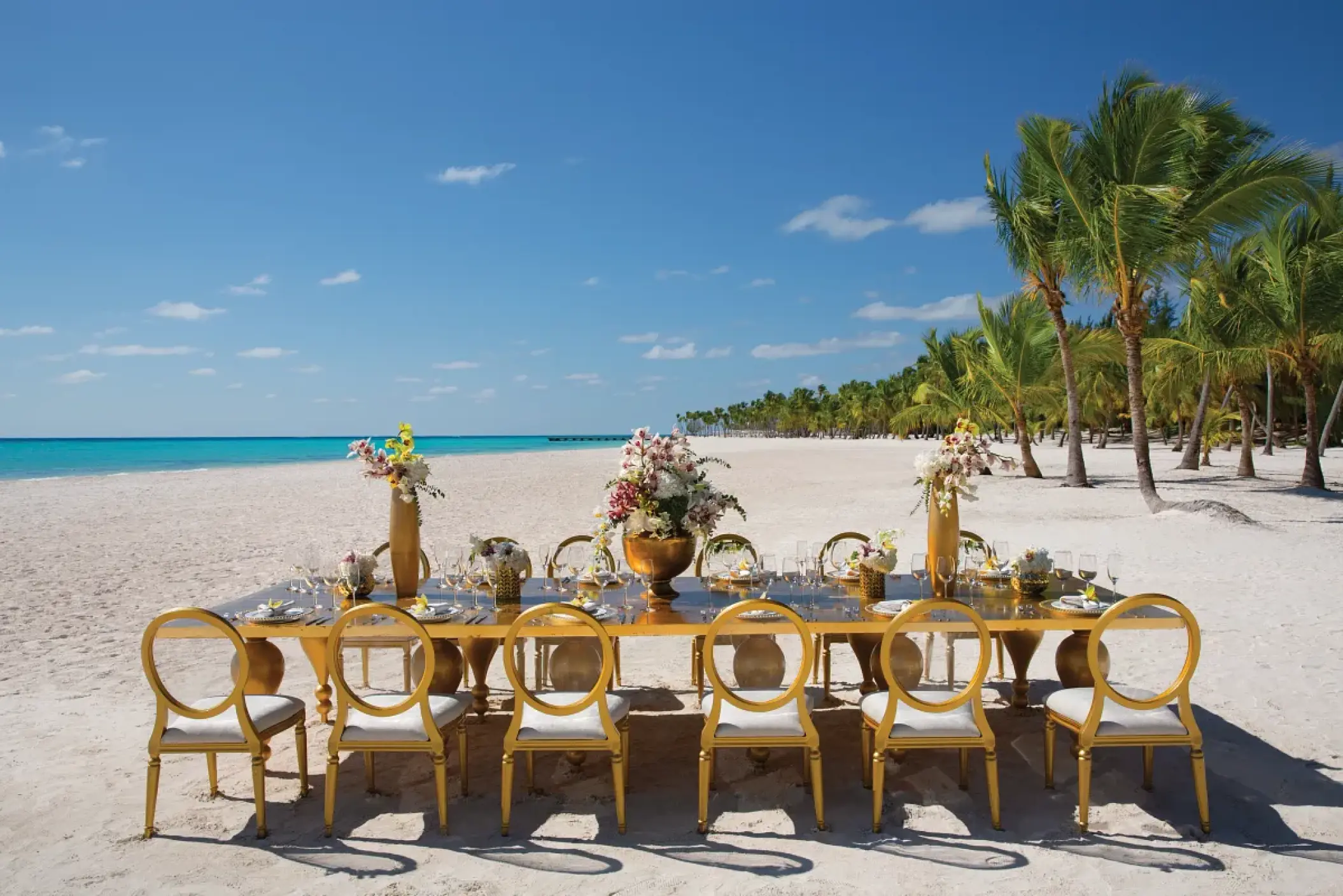 Long gold reception table with floral centerpieces on the beach at Secrets Cap Cana, Punta Cana