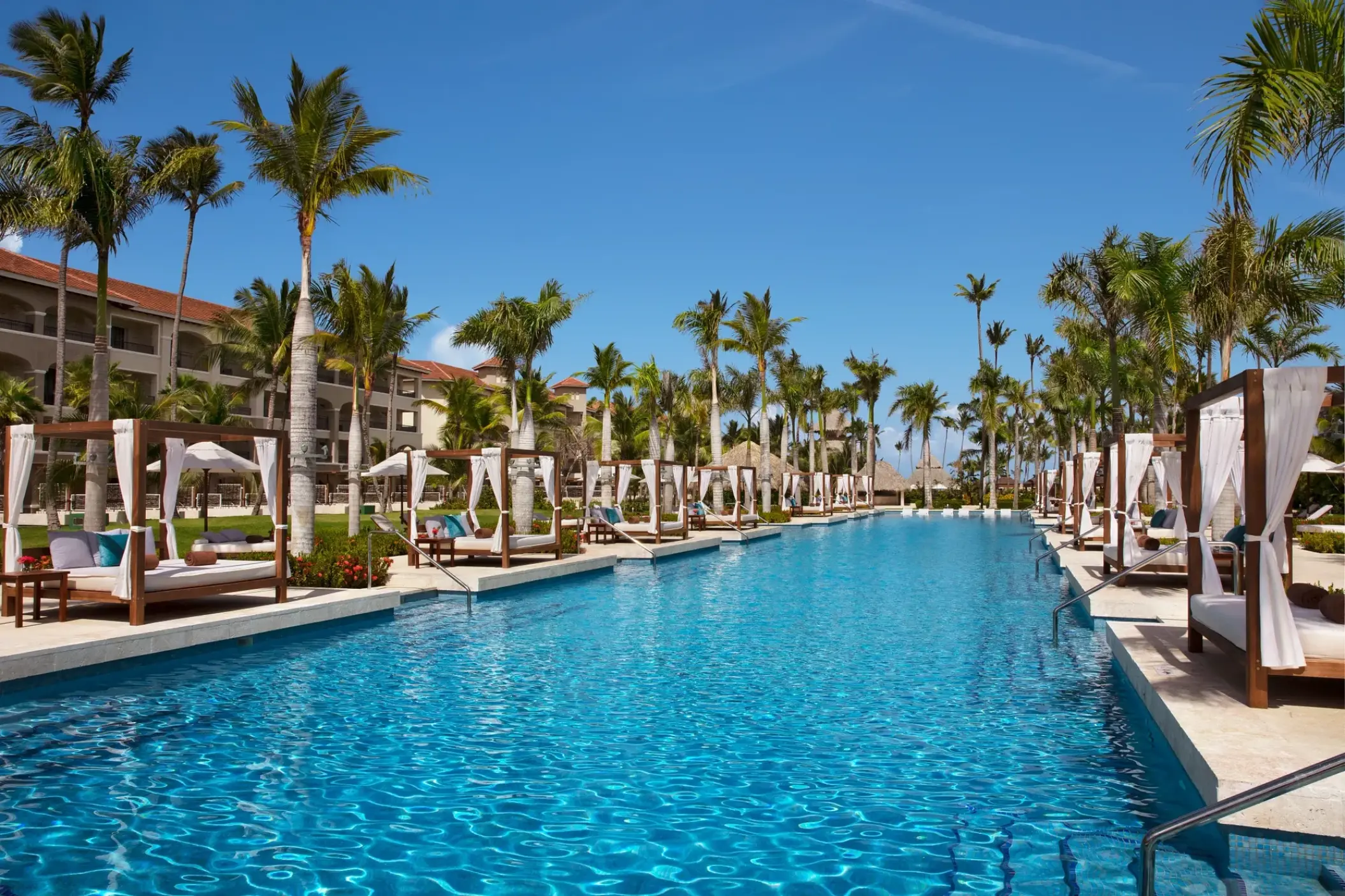 Long blue swimming pool lined with white cabanas and palm trees at Secrets Royal Beach, Punta Cana
