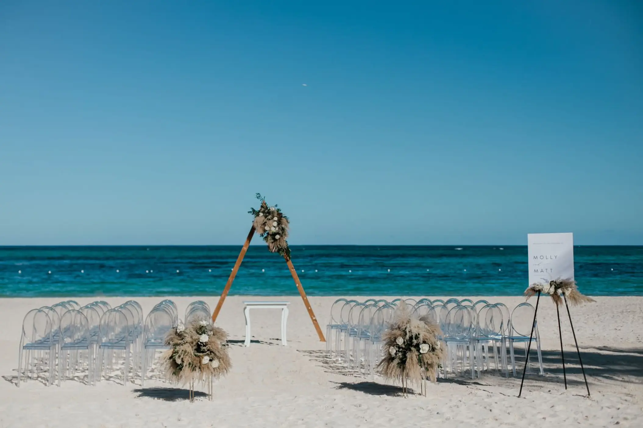 Beachfront wedding ceremony with a triangular wooden arch and acrylic chairs at Secrets Royal Beach, Punta Cana