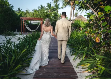 Danielle & Derrek walking hand-in-hand along a tropical wooden boardwalk toward the beach