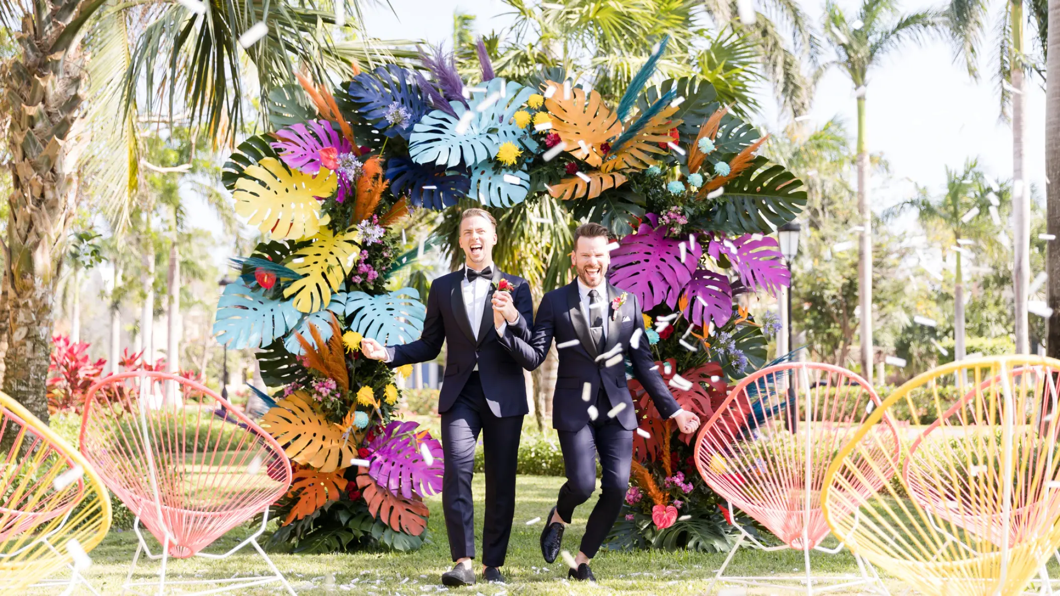 Two grooms celebrating under a colorful monstera leaf arch at Iberostar Selection Cancun