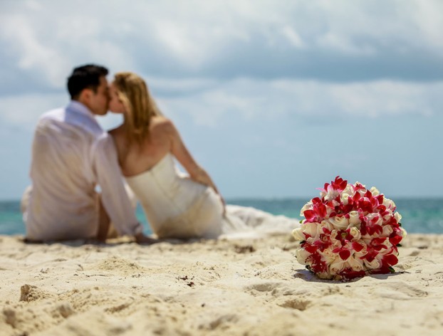 Couple kisses on the beach on the background as the bride's bouquet shows in foreground.