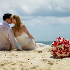 Couple kisses on the beach on the background as the bride's bouquet shows in foreground.