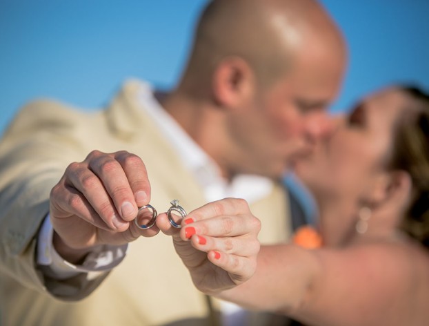 Bride and Groom showing their wedding bands while kissing on background.
