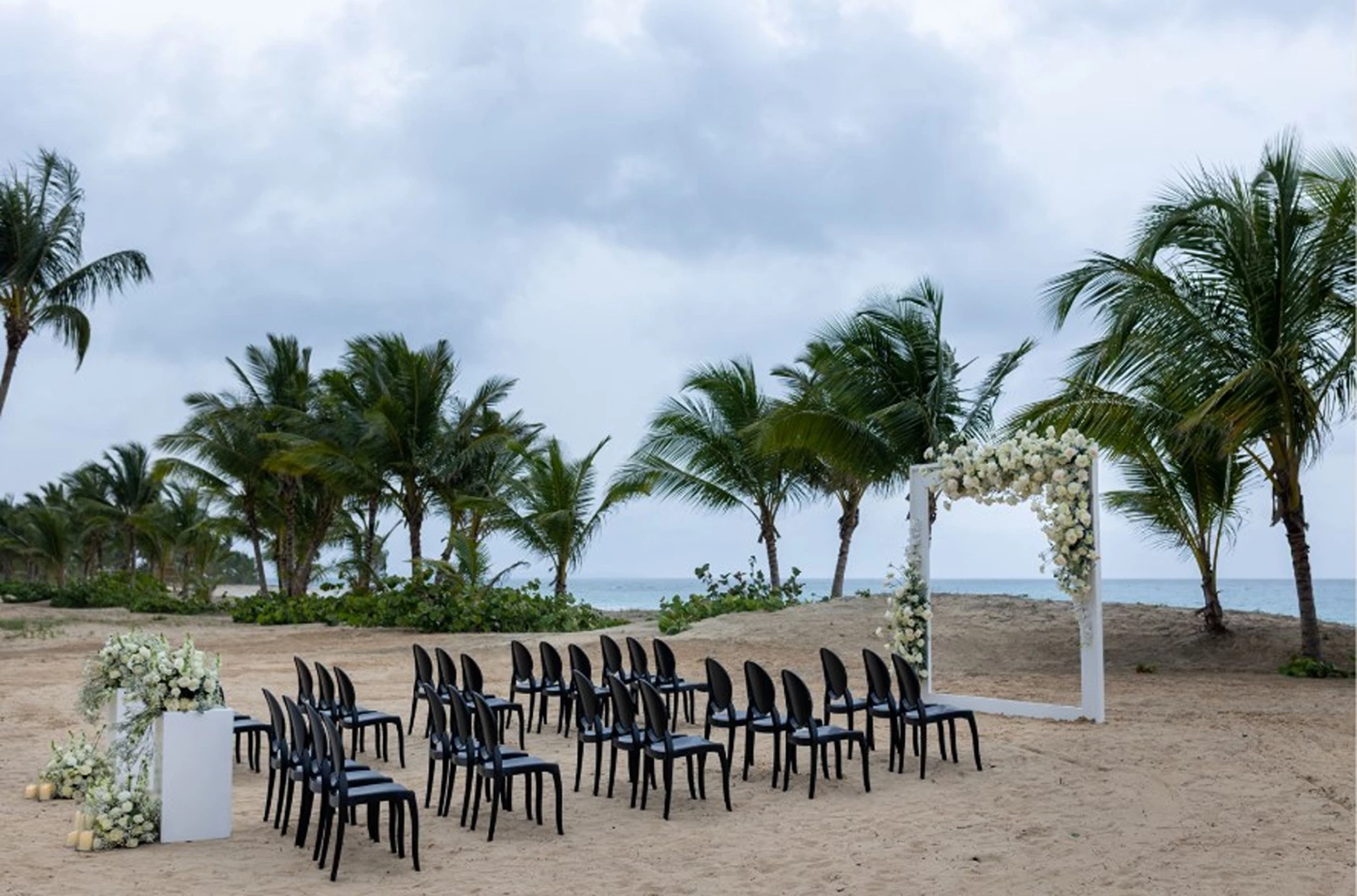A beach wedding setup with rows of black chairs facing a floral arch, with the ocean in the background.