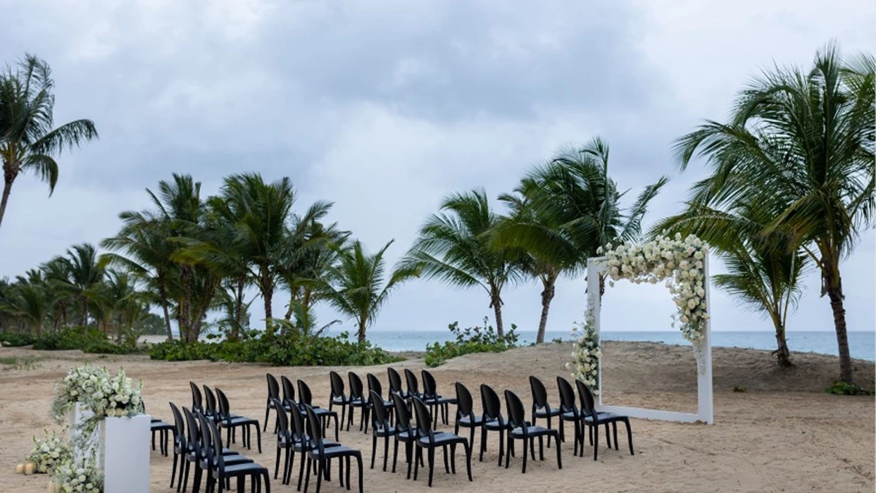 A beach wedding setup with rows of black chairs facing a floral arch, with the ocean in the background.