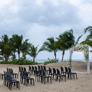 A beach wedding setup with rows of black chairs facing a floral arch, with the ocean in the background.