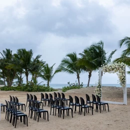 A beach wedding setup with rows of black chairs facing a floral arch, with the ocean in the background.