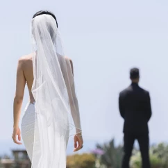 A bride in a flowing white dress and veil walks towards the groom, who is out of focus in the background.