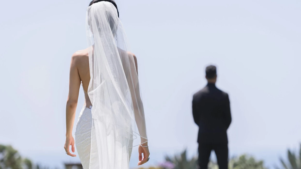 A bride in a flowing white dress and veil walks towards the groom, who is out of focus in the background.
