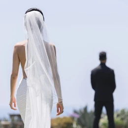 A bride in a flowing white dress and veil walks towards the groom, who is out of focus in the background.