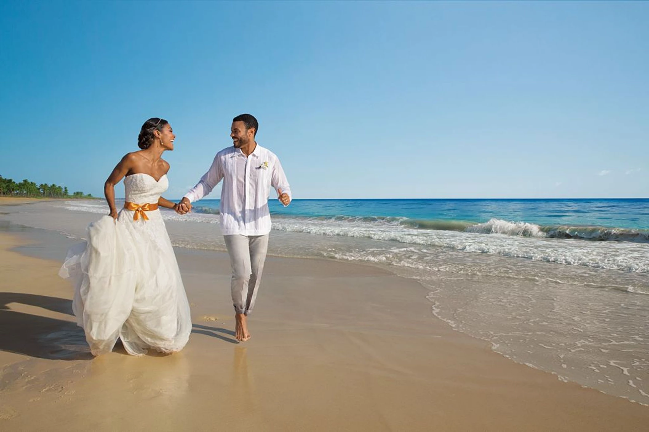 A joyful bride and groom, in wedding attire, holding hands and running along the beautiful sandy beach.