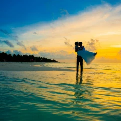 A silhouetted bride and groom embrace in the shallow ocean water during a vibrant, colorful sunset.