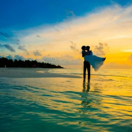 A silhouetted bride and groom embrace in the shallow ocean water during a vibrant, colorful sunset.