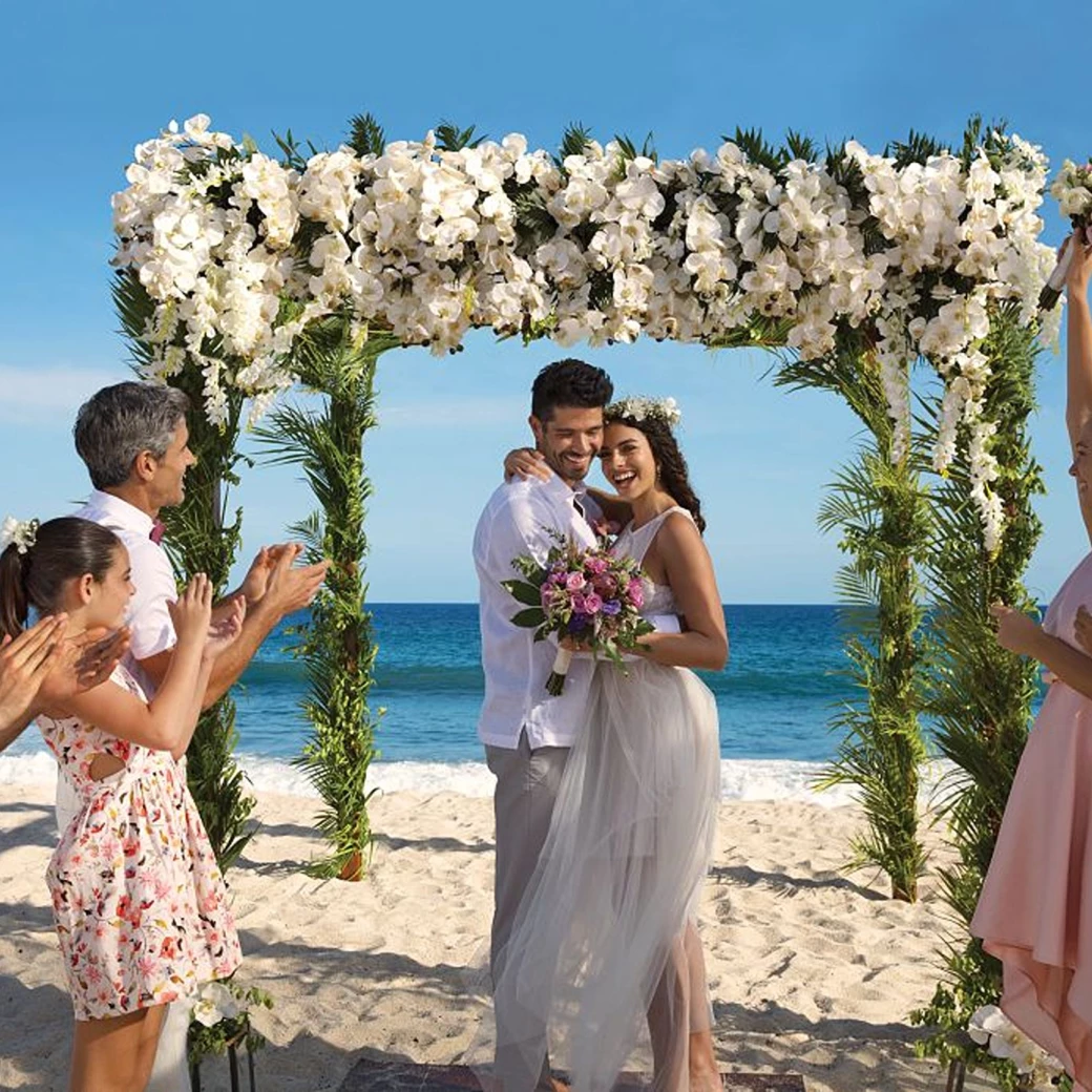 A newly married couple and their happy guests celebrating on a sunlit beach under a beautiful floral arch.