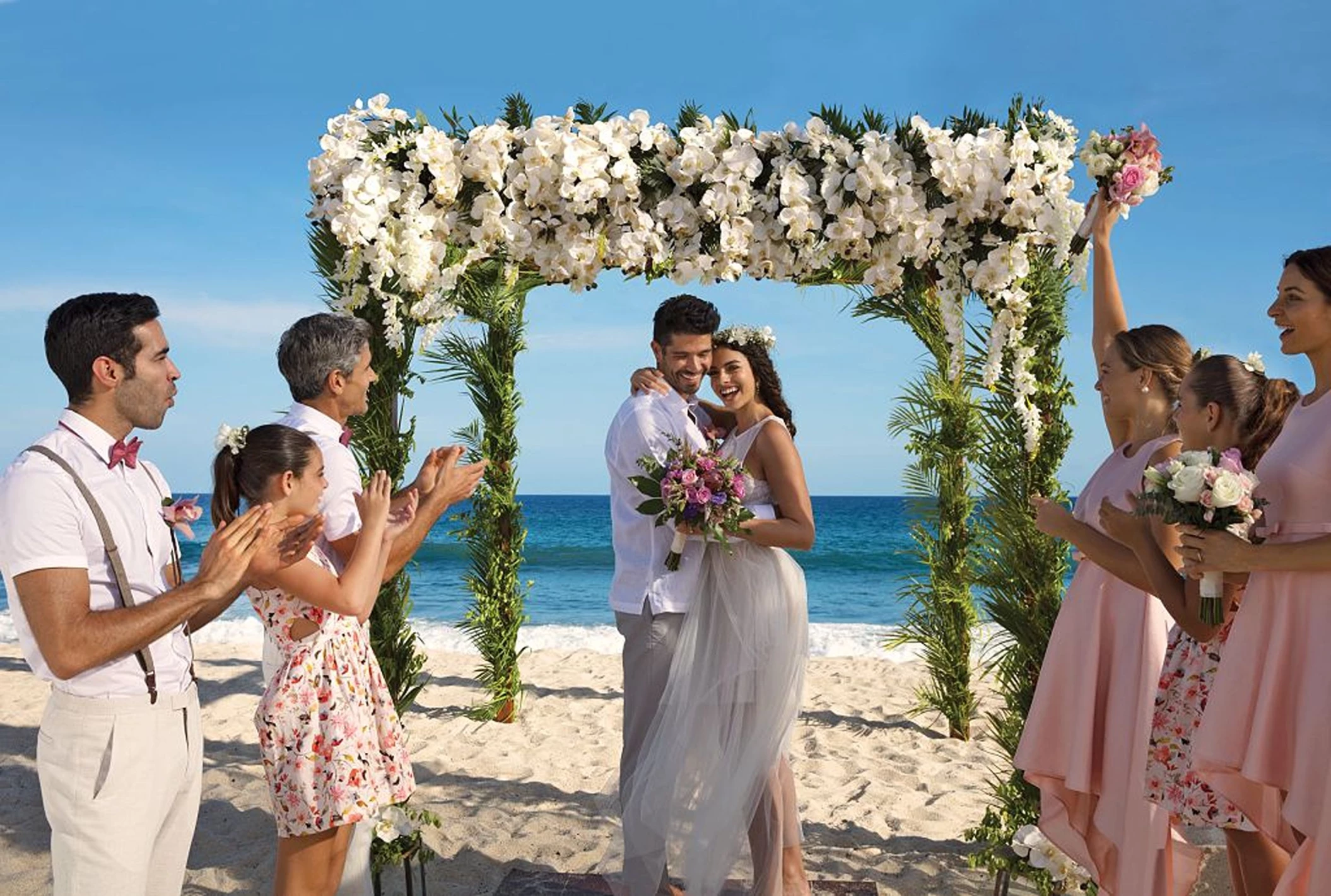 A newly married couple and their happy guests celebrating on a sunlit beach under a beautiful floral arch.