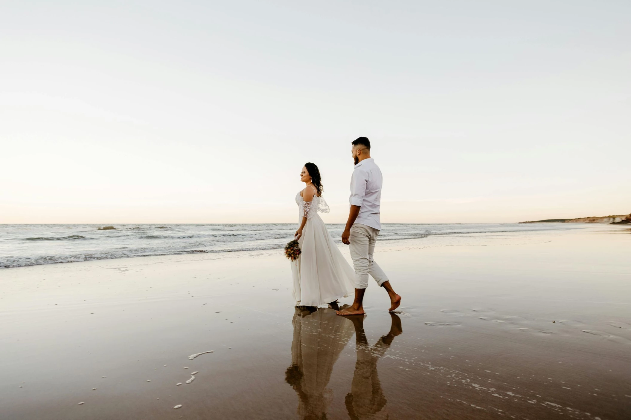 A bride and groom walking barefoot on a wet, sandy beach, their reflections visible in the water.