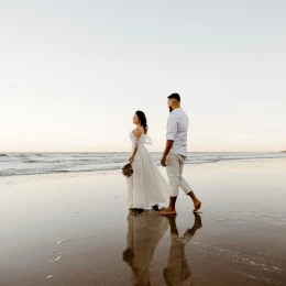 A bride and groom walking barefoot on a wet, sandy beach, their reflections visible in the water.
