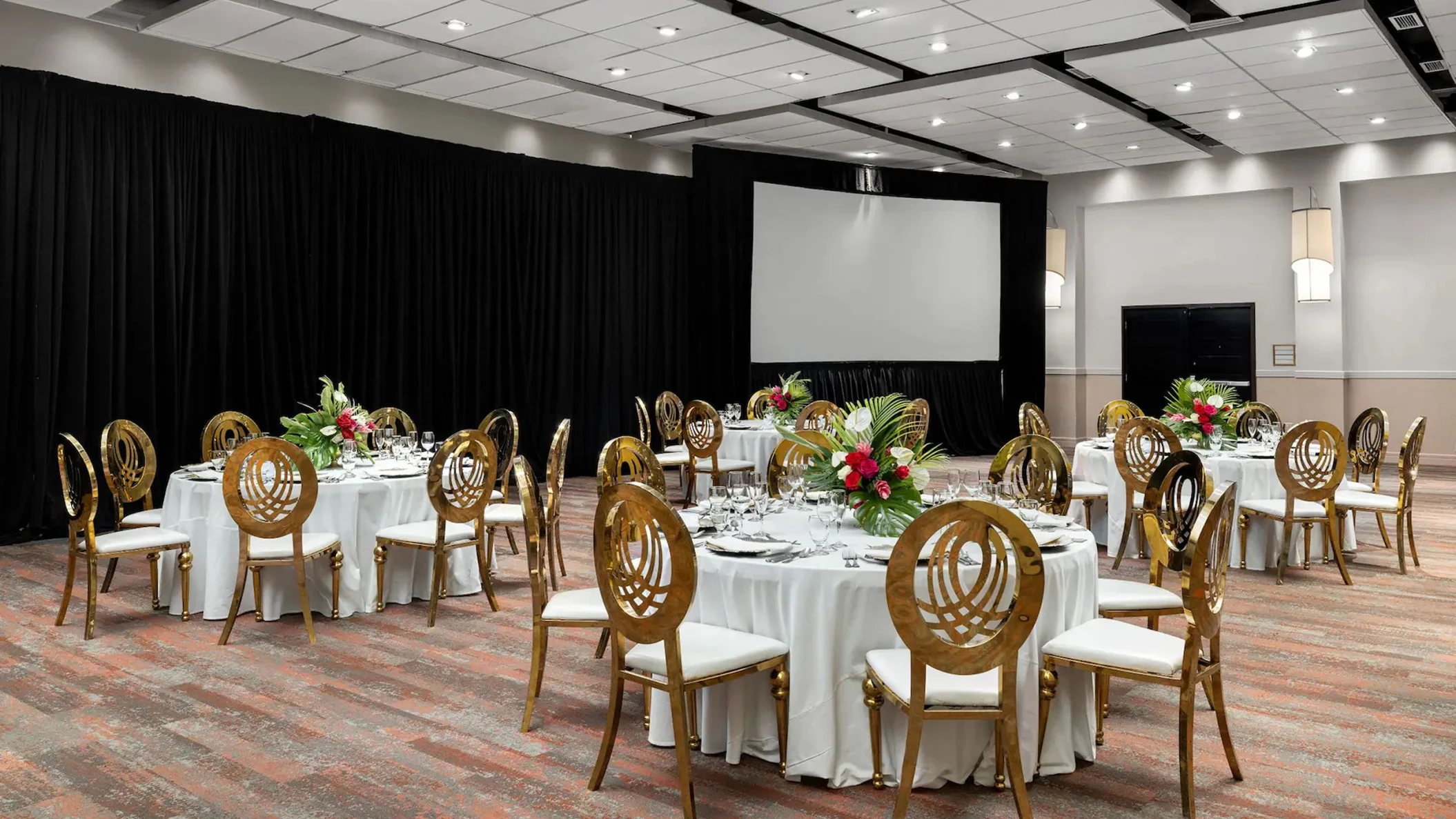The beautifully set-up ballroom venue at the resort, featuring round tables, elegant gold chairs, and a screen.