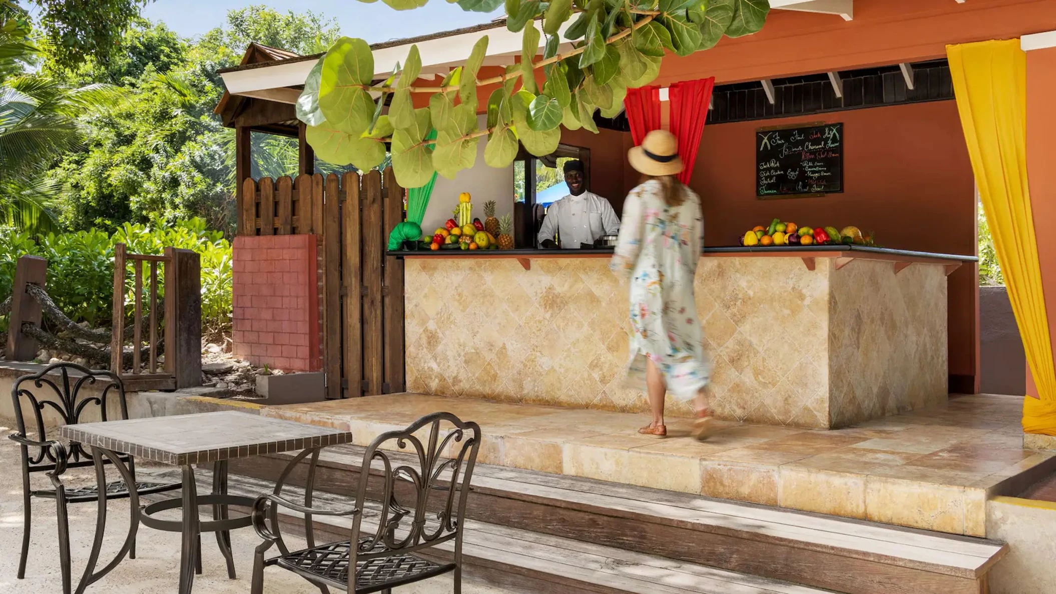 The resort's outdoor Jerk Hut with a bartender, a guest, and a selection of fresh fruits on the counter.