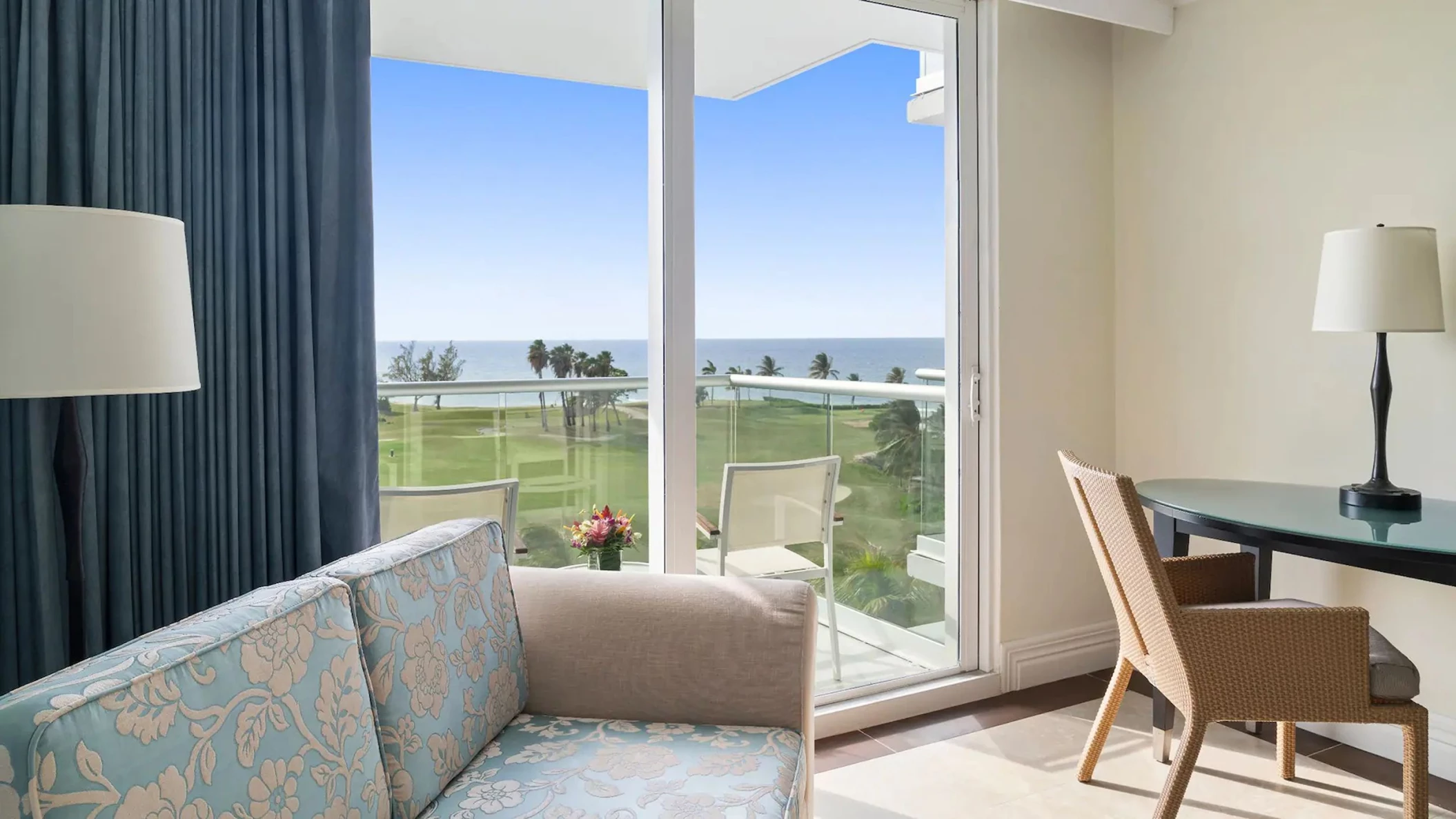 Living area of a Partial Ocean View King room with a sofa and desk, looking out onto a balcony and the ocean.