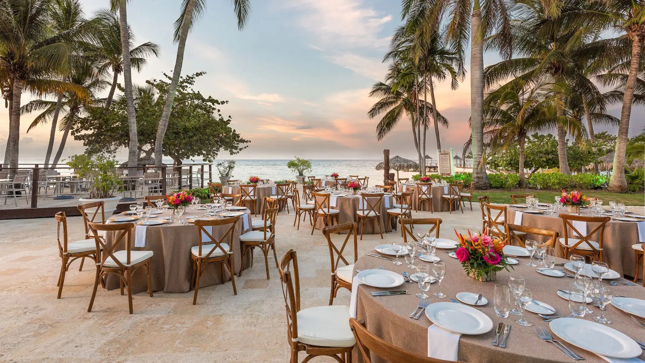An outdoor wedding reception at Mangoes, with tables and chairs set up under palm trees by the beach at sunset.