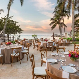 An outdoor wedding reception at Mangoes, with tables and chairs set up under palm trees by the beach at sunset.
