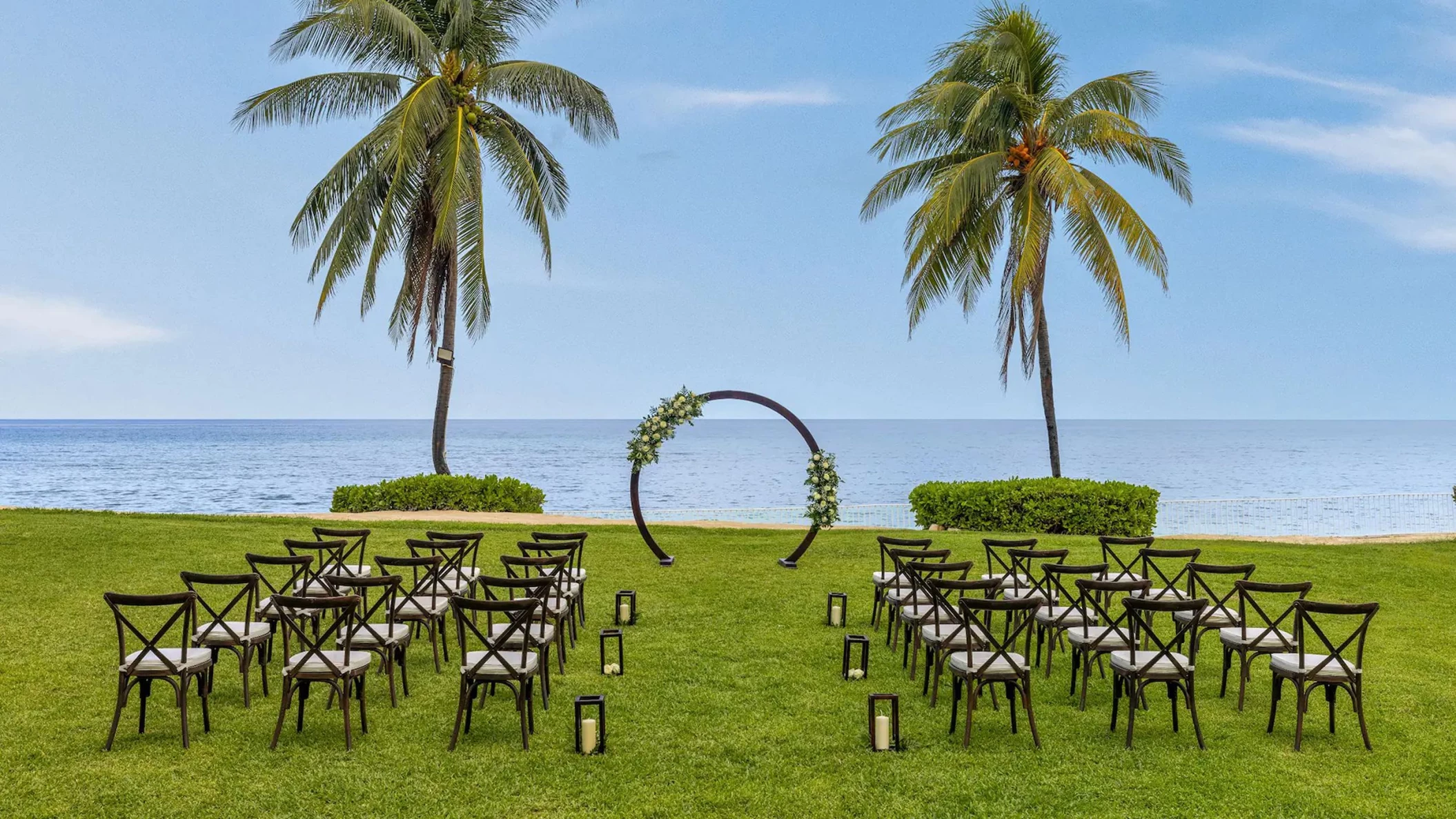 An outdoor wedding venue on the West Lawn with an arch, chairs, and lanterns set up with the ocean as the backdrop.