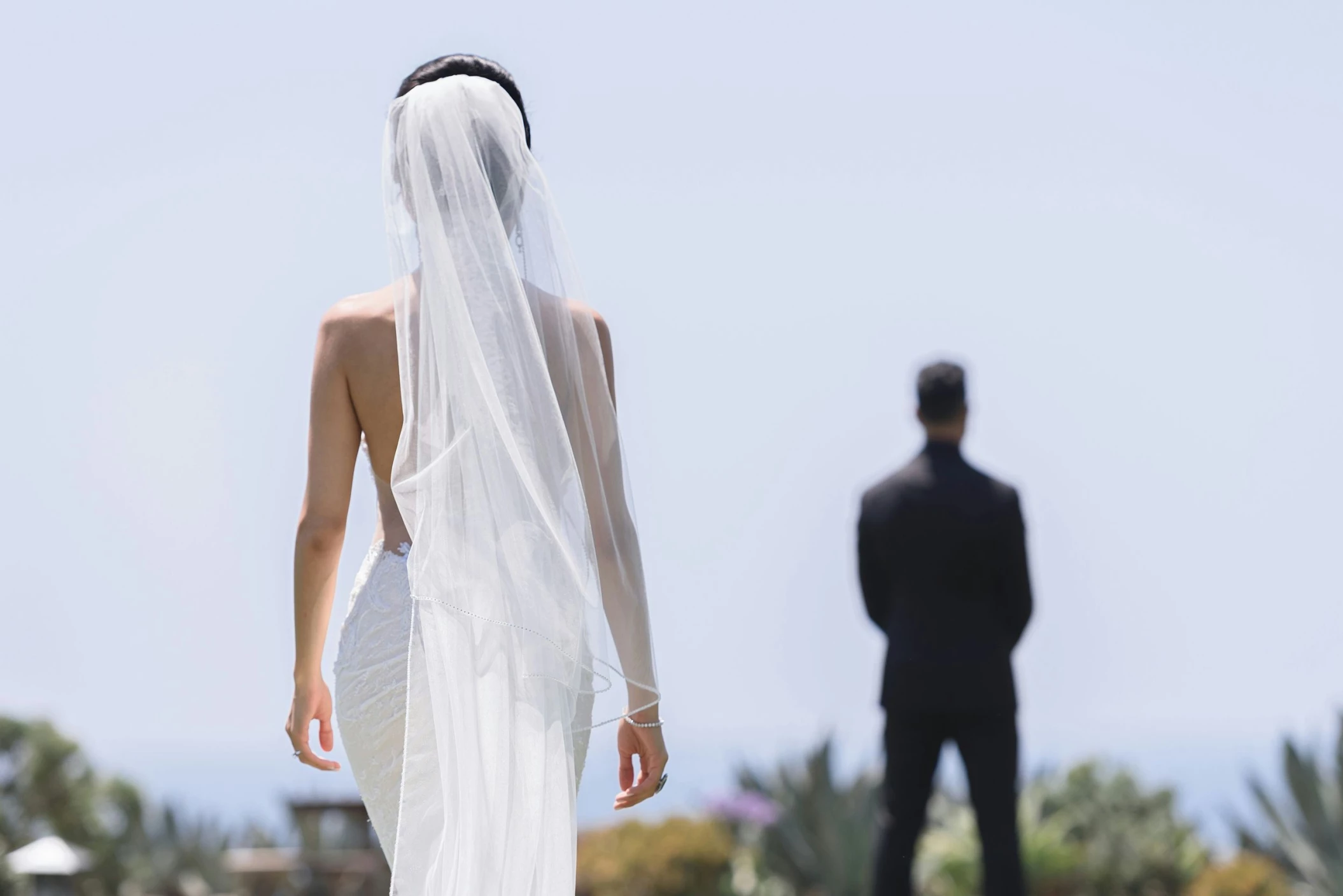 A bride in a flowing white dress and veil walks towards the groom, who is out of focus in the background.