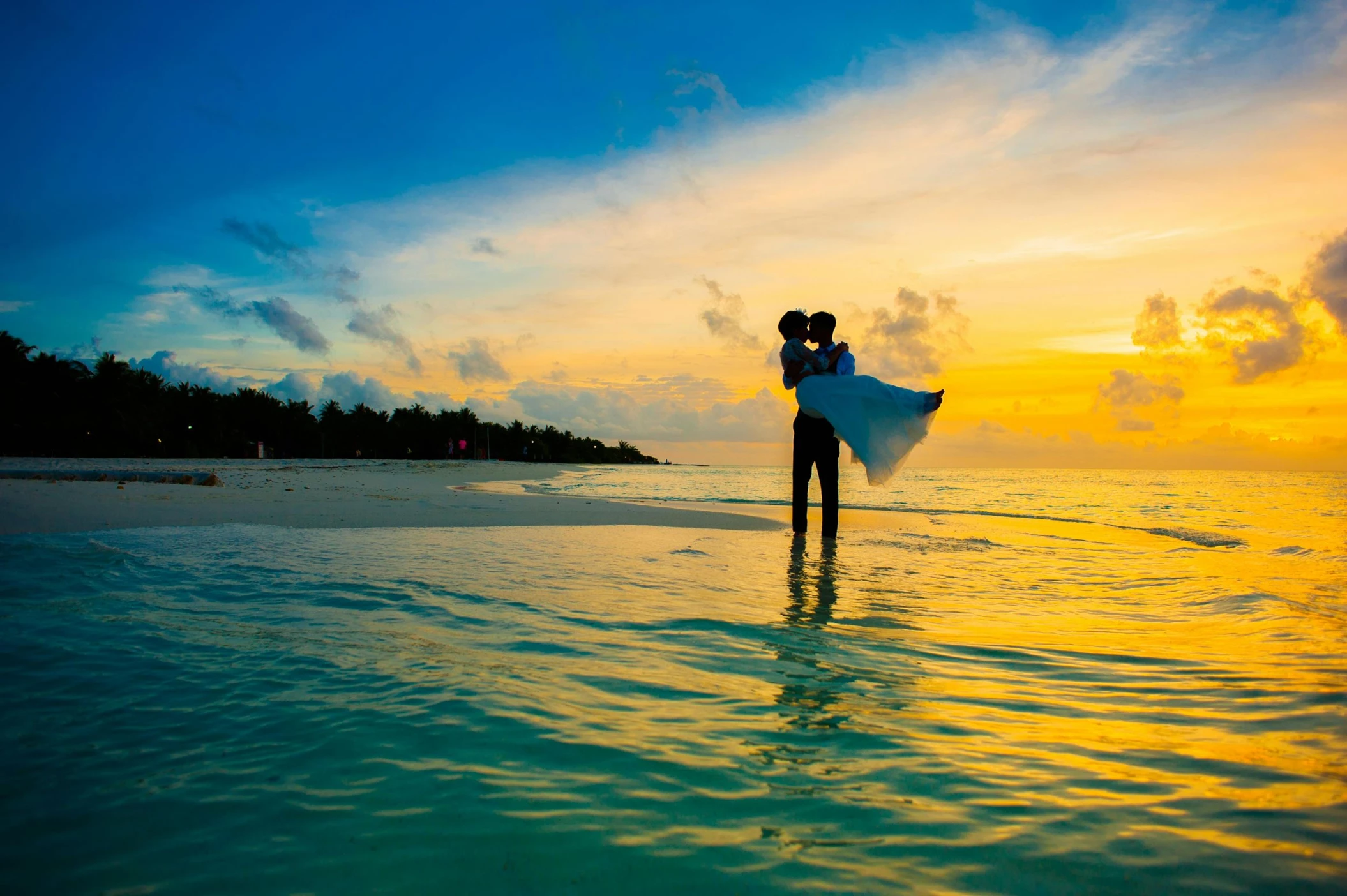 A silhouetted bride and groom embrace in the shallow ocean water during a vibrant, colorful sunset.