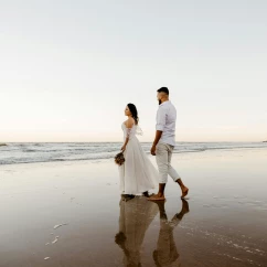 A bride and groom walking barefoot on a wet, sandy beach, their reflections visible in the water.