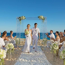 A happy couple walking down a petal-strewn aisle on the beach, surrounded by applauding guests.