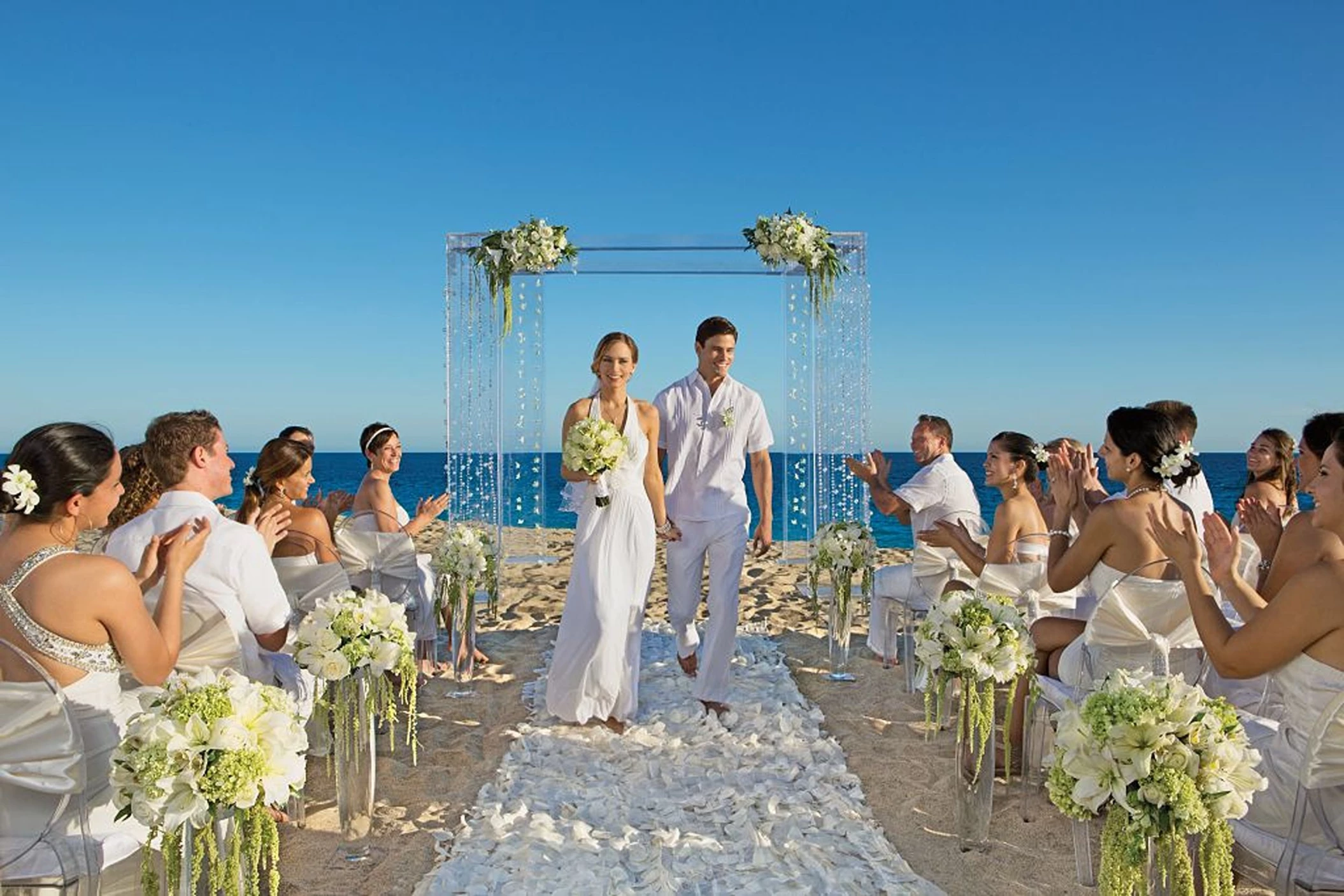 A happy couple walking down a petal-strewn aisle on the beach, surrounded by applauding guests.
