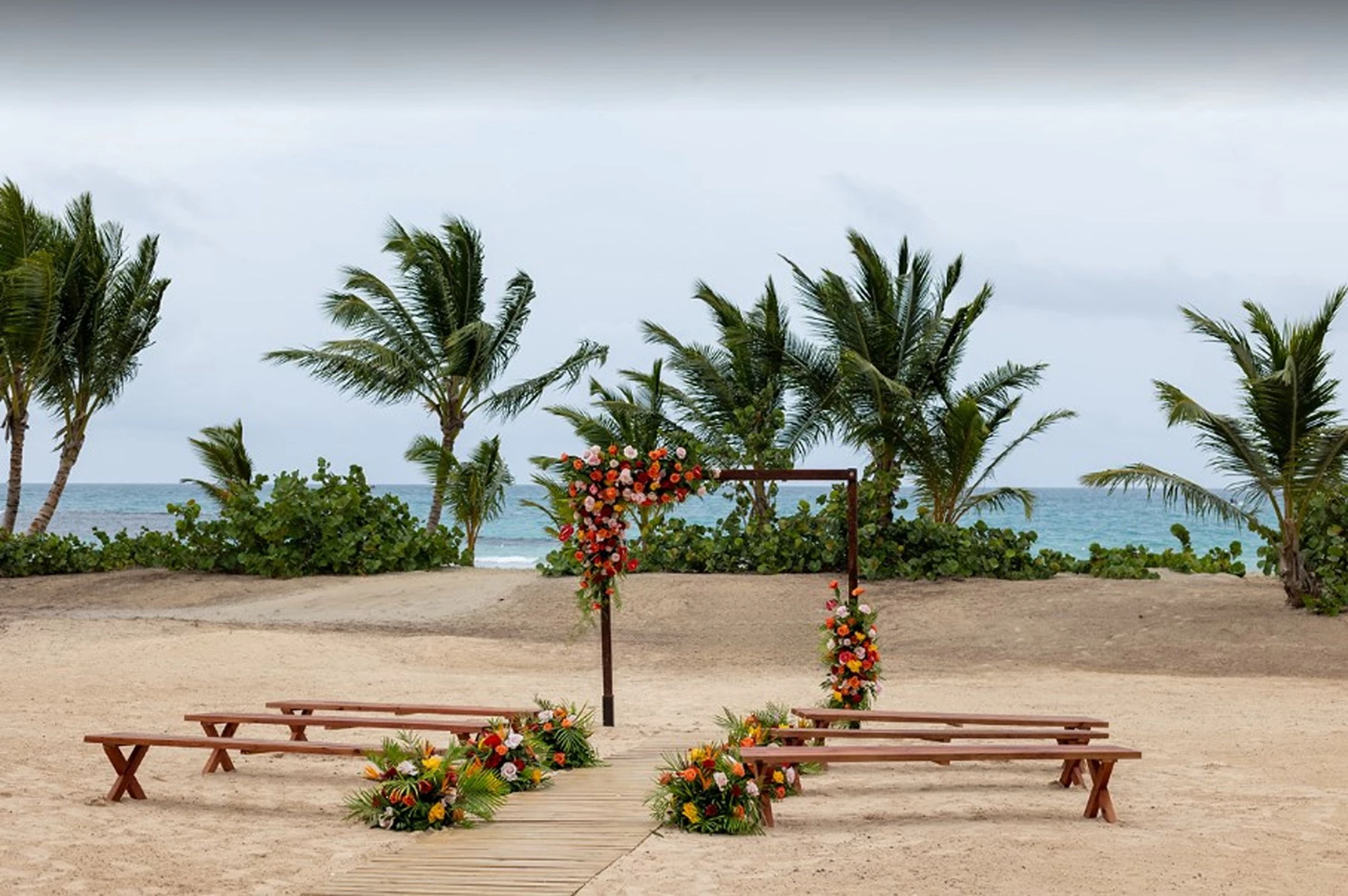 A beach wedding venue with a wooden walkway leading to a floral arch, with benches and palm trees.