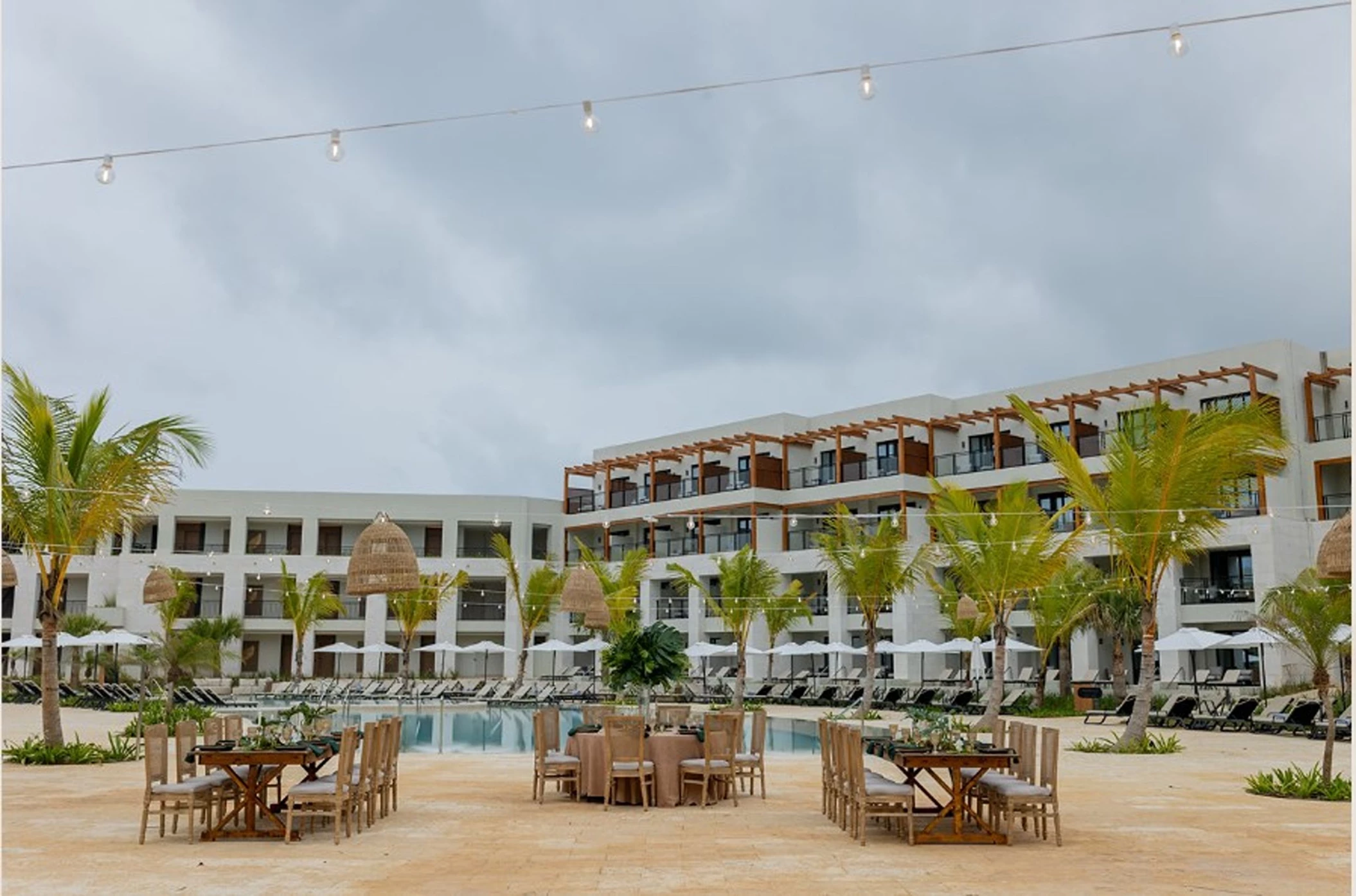 An outdoor dining setup with tables and chairs on a terrace, overlooking a large swimming pool and hotel.