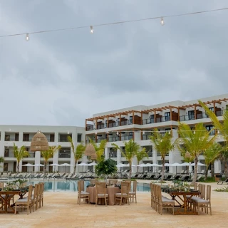 An outdoor dining setup with tables and chairs on a terrace, overlooking a large swimming pool and hotel.