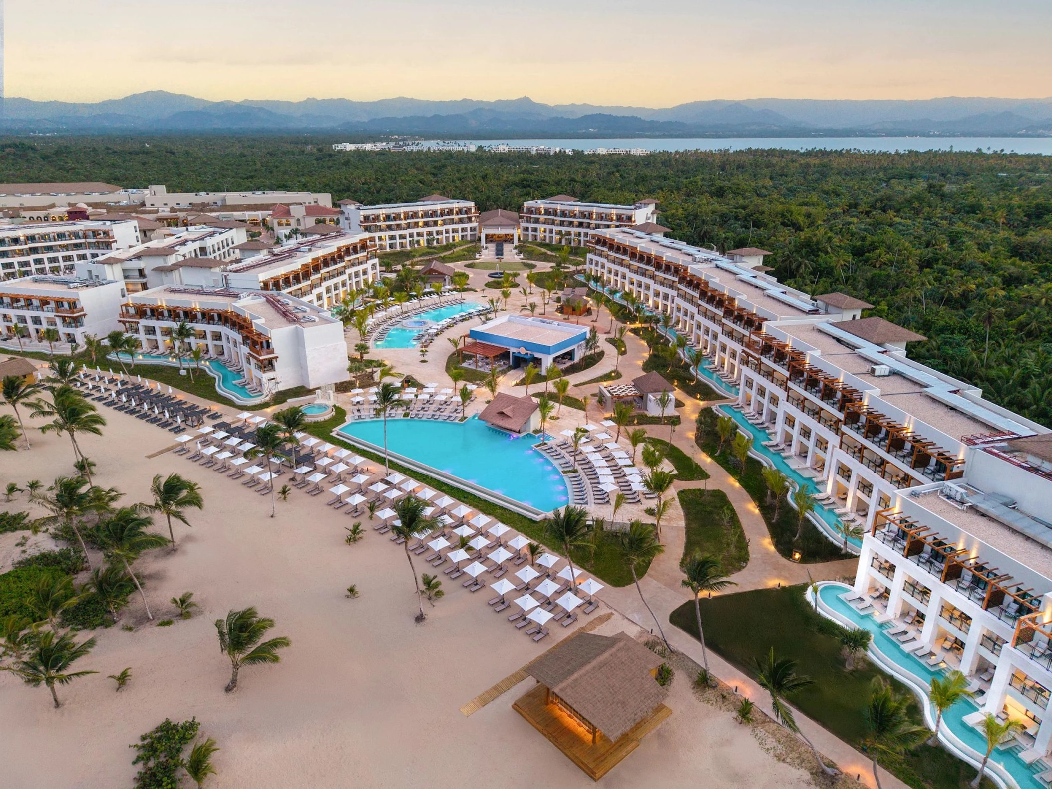 An aerial view of the Secrets Playa Esmeralda resort at sunset, with pools, beach, and lush trees.
